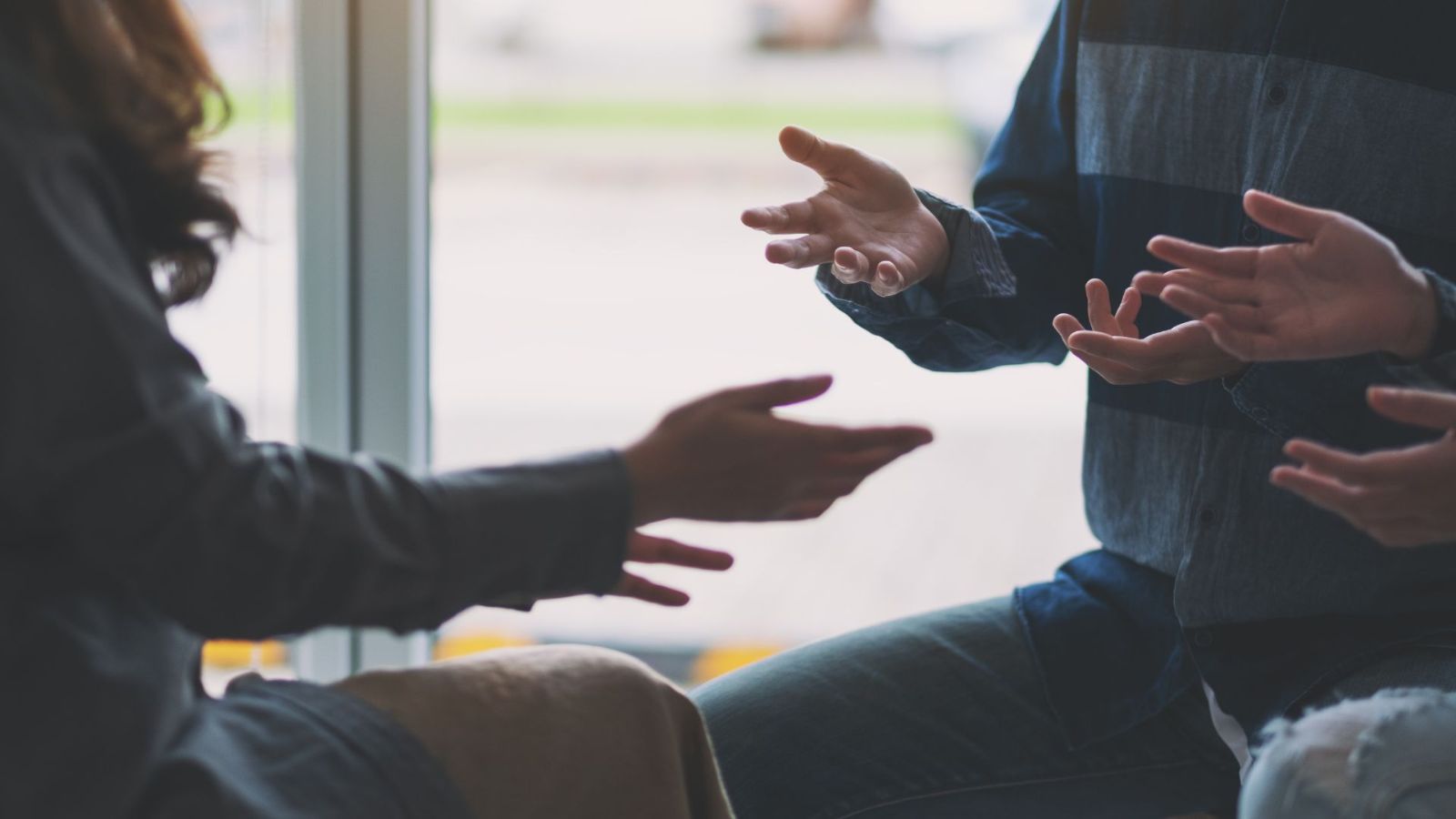 Close de mãos de duas pessoas em conversa, gesticulando junto a uma janela